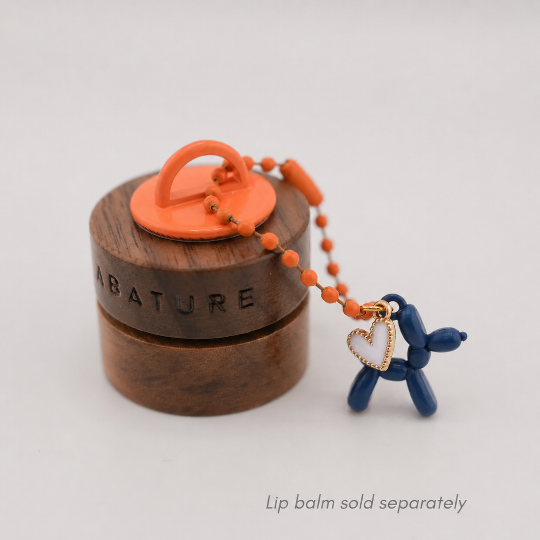 A small round wooden lip balm container with an orange lid and handle, shown on a light gray background. A beaded orange chain hangs from the lid and includes a blue balloon-style dog charm with a gold heart pendant.
